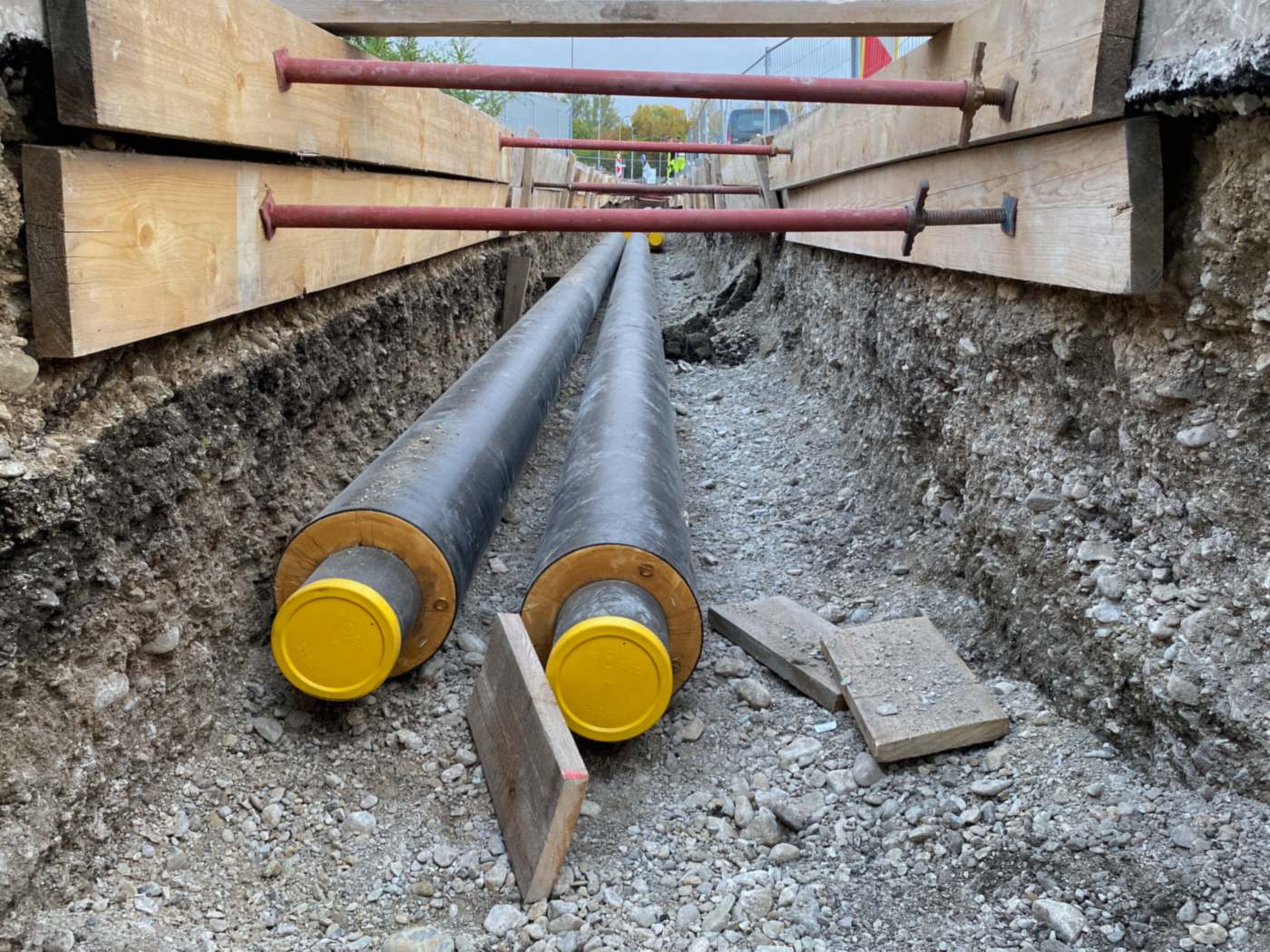 A shored utility trench: timber sheeting along the upper walls is held apart by red steel trench struts, and two black pre-insulated pipes with yellow end caps lie on a gravel bed. Loose boards lie on the trench bottom.