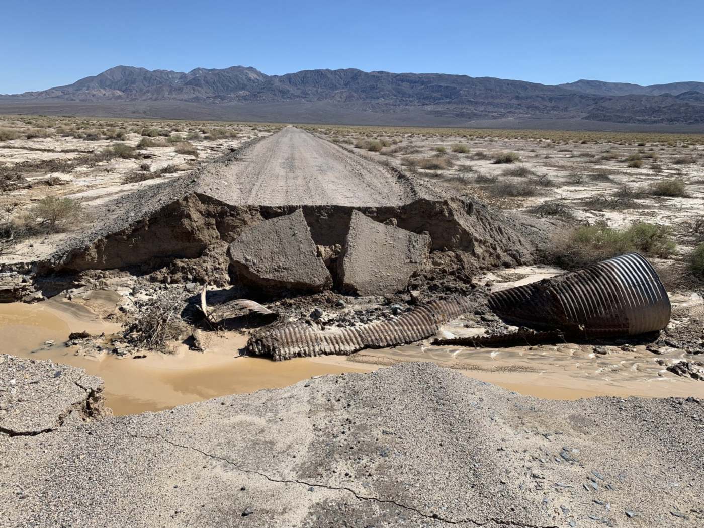 A desert road washed out by floodwater. The roadway ends in a broken edge, two large asphalt slabs have dropped into the gap, and a corrugated metal culvert pipe lies crushed and torn in muddy water below. Woody debris sits in the mud, and the road continues towards mountains on the far side.