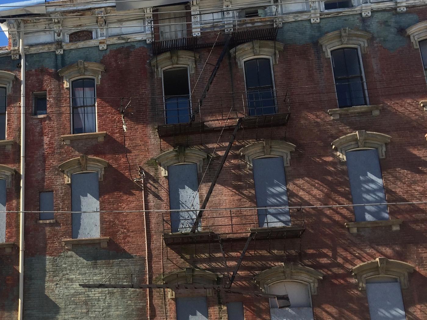 Close-up of a weathered four-story brick facade: a painted cornice with peeling paint and a damaged bracket, carved window hoods, boarded window openings, painted brick with paint loss, and a rusted steel fire escape with a counterbalanced stair.