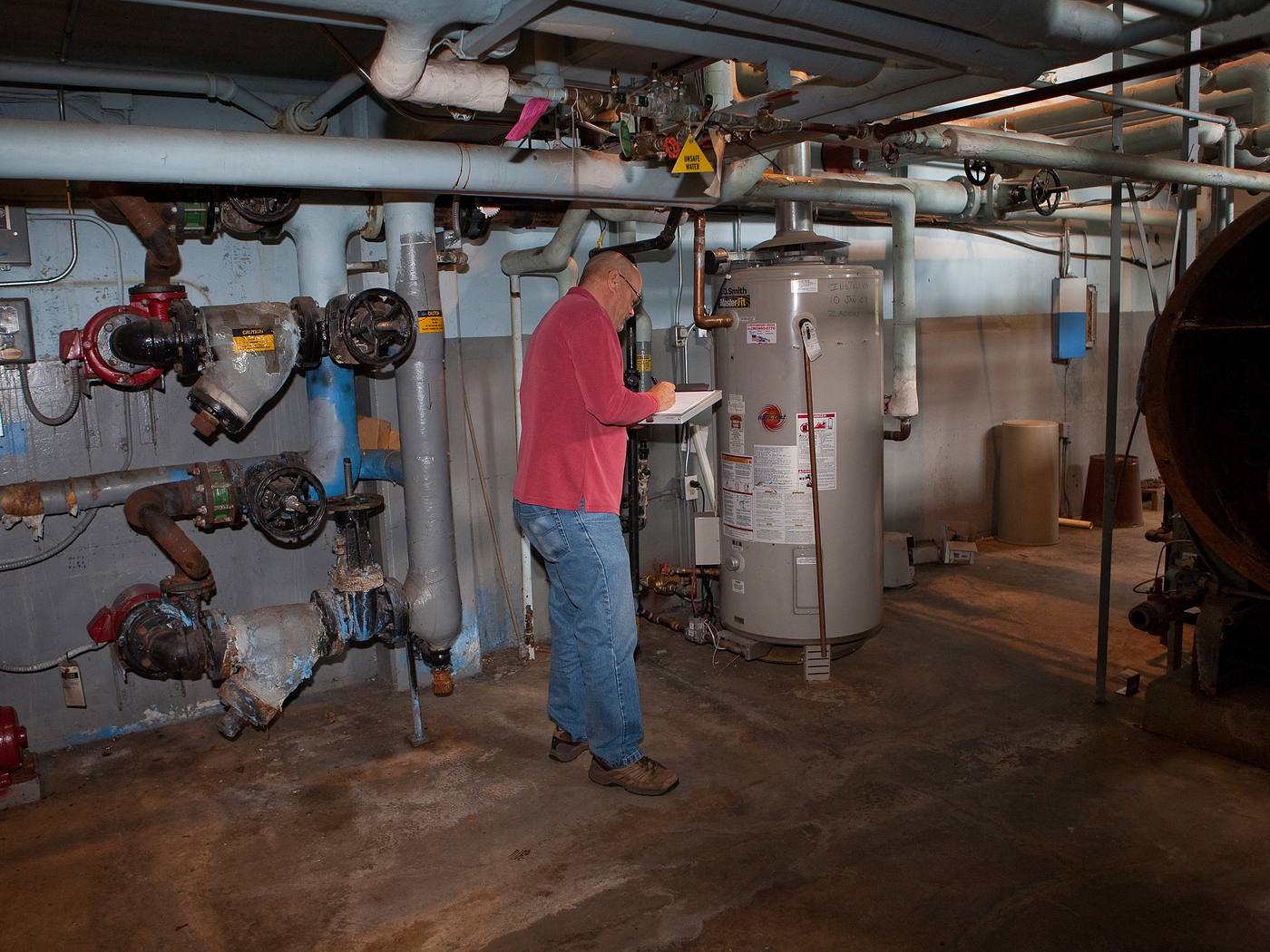 An assessor with a clipboard records data beside a gas water heater in a school mechanical room. On the wall to the left, a red circulator pump, valves and a pipe strainer are heavily corroded, and the insulation on overhead pipe fittings is torn. A rusted boiler shell stands at the right edge, and the concrete floor is stained.
