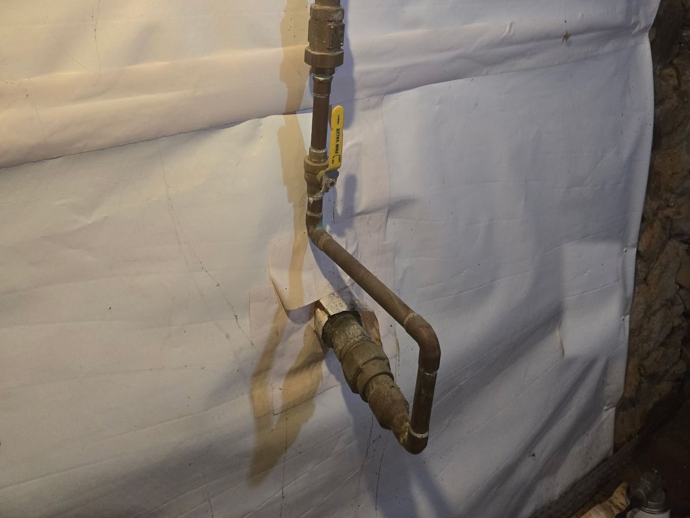 A water service pipe enters a basement through the foundation wall into a brass coupling. Copper piping bends up from the coupling to a ball valve with a yellow handle, against a white vapor barrier.