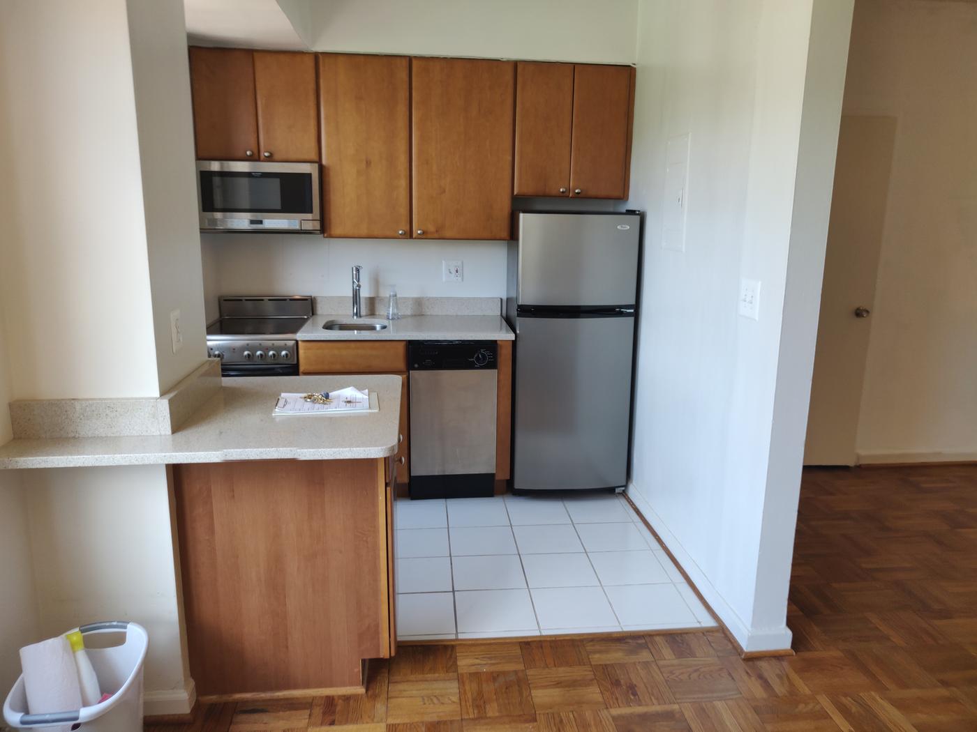 A vacant apartment kitchen with wood cabinets, an over-range microwave, an electric range, a small sink with an outlet beside it, a dishwasher and a stainless refrigerator on white floor tile. An electrical panel cover and a light switch are on the right-hand wall, and a raised wood strip separates the tile from the parquet floor. A clipboard with keys lies on the counter.