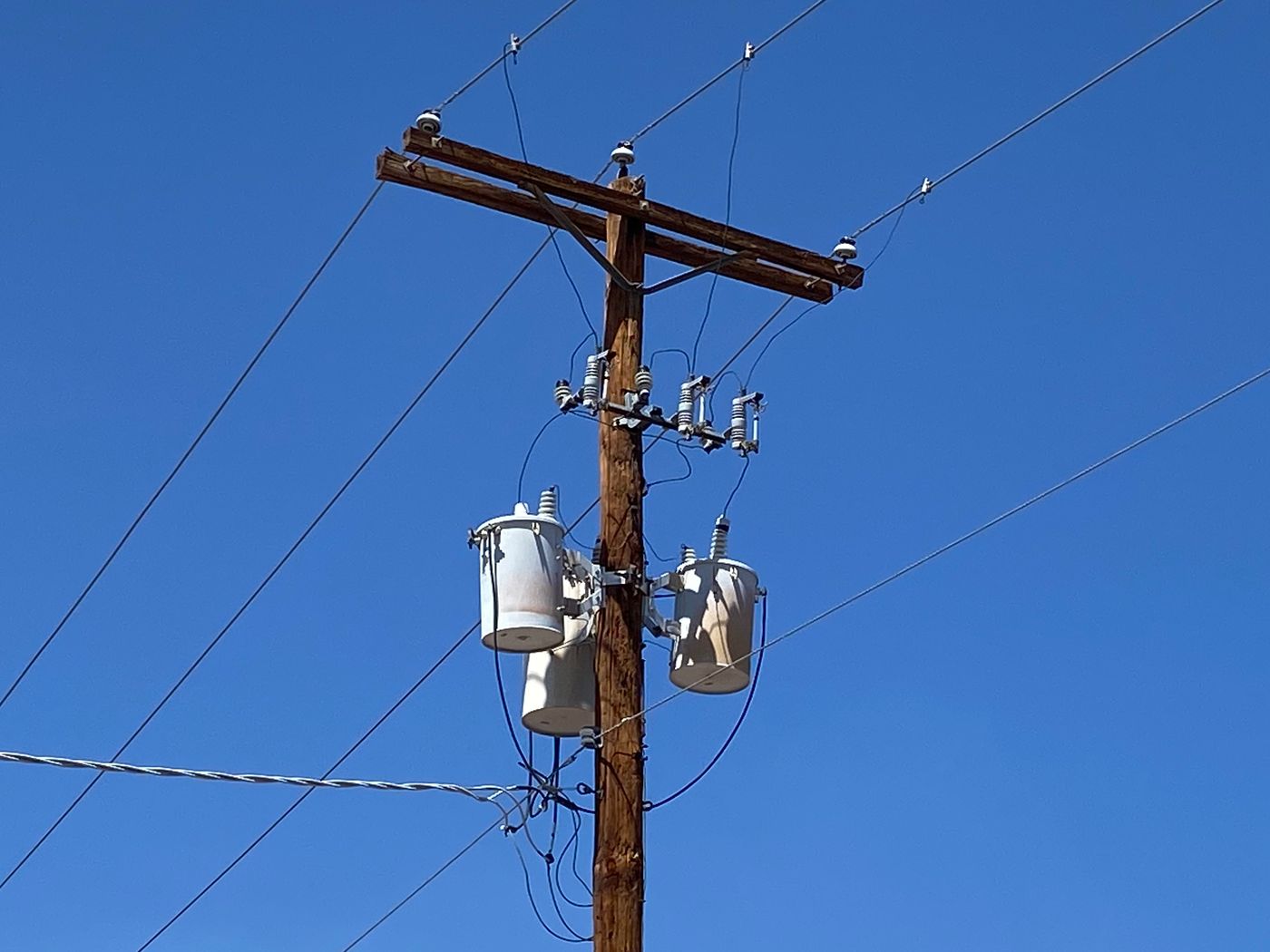 A wood distribution pole against a clear blue sky: a double crossarm with pin insulators at the top, a row of fused cutouts below it, a bank of three pole-mounted transformers, and a twisted service drop attached lower on the pole.