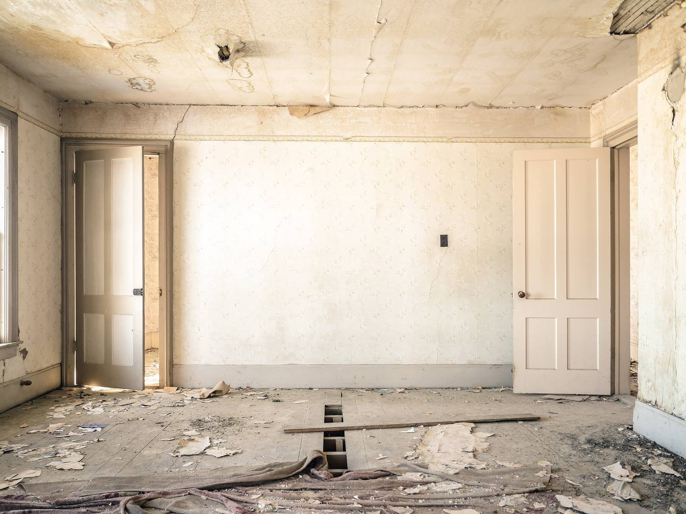 An empty room in an older house: a water-stained ceiling with a hole and a long crack, a diagonal crack running up from the head of the left-hand door, a hairline crack below the wall outlet, a vertical crack with lost plaster on the right-hand wall, and missing floorboards in the middle of the floor.