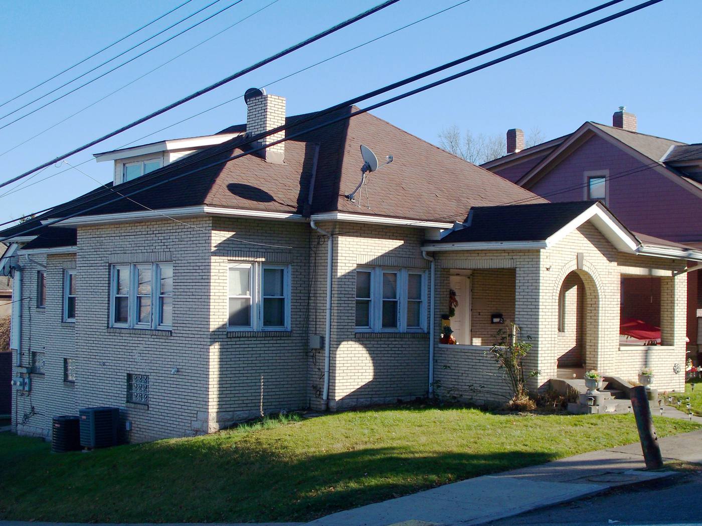 A one-storey brick house with a brown asphalt-shingle hip roof, a dormer and a brick chimney with a satellite dish on top. Overhead power lines cross the frame above the roof. A small arched porch at the right has concrete steps with no handrail.