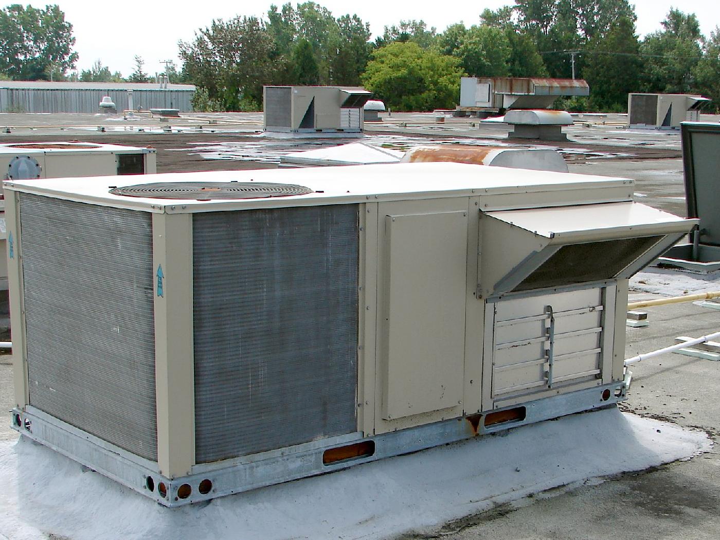 A packaged rooftop HVAC unit sits on a coated curb on a low-slope commercial roof. Behind it are wet patches of standing water, more rooftop units, exhaust fans and a rusted section of ductwork. Gas and condensate lines run on pipe supports at the right.