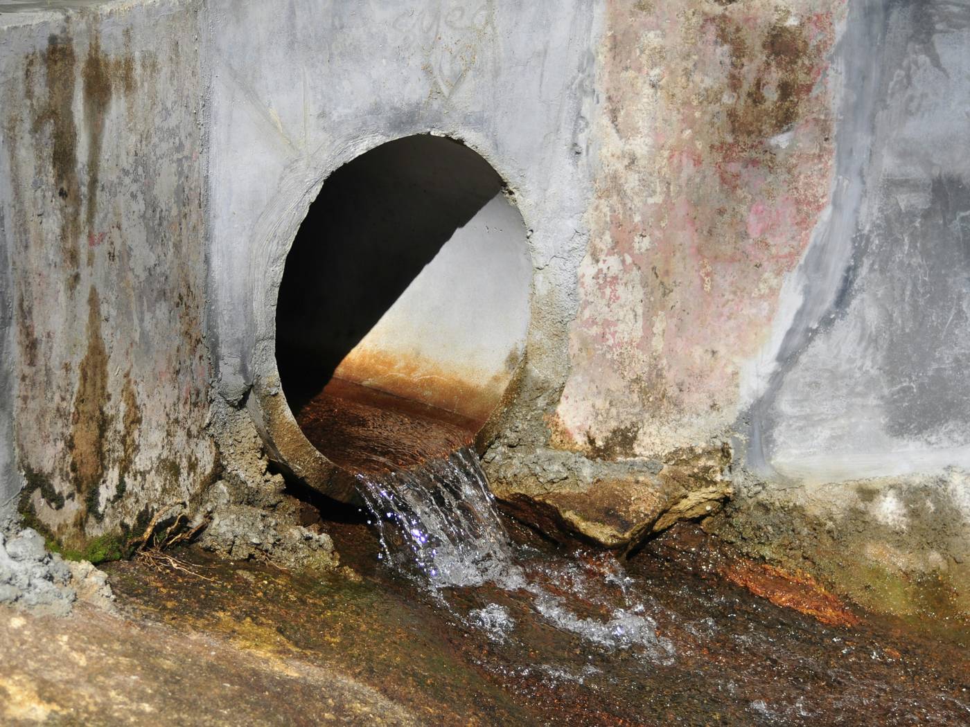 A round concrete pipe outfall set in a stained concrete headwall. A thin stream of clear water falls from the pipe onto a channel bed with orange-brown deposits. The pipe invert is stained rust-orange and the concrete at the outlet is broken.