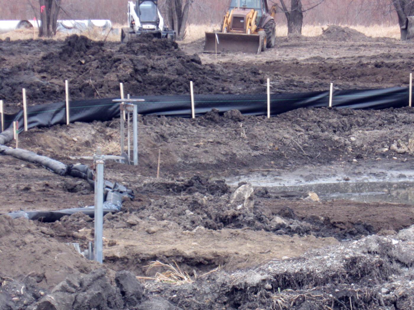 A muddy construction site. A black silt fence on wooden stakes runs across the middle, with a soil stockpile, a skid steer and a backhoe behind it. In front, a black filter sock lies kinked on bare soil beside metal posts, and a shallow pool of muddy water stands on the right.